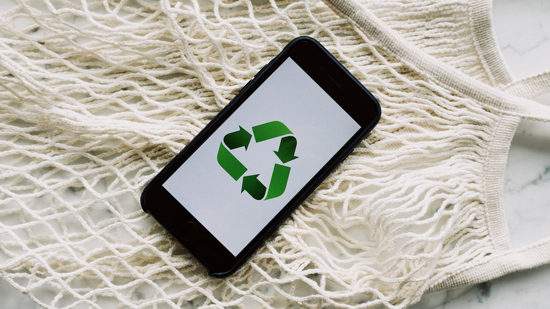 A phone with the green recycle logo on it's screen placed on top of a marble counter and string grocery bag